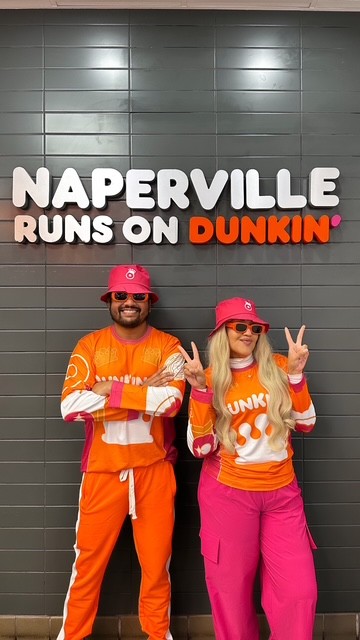 Two PPP Donuts Network crew members in Dunkin' gear in front of a 'Naperville Runs on Dunkin' wall