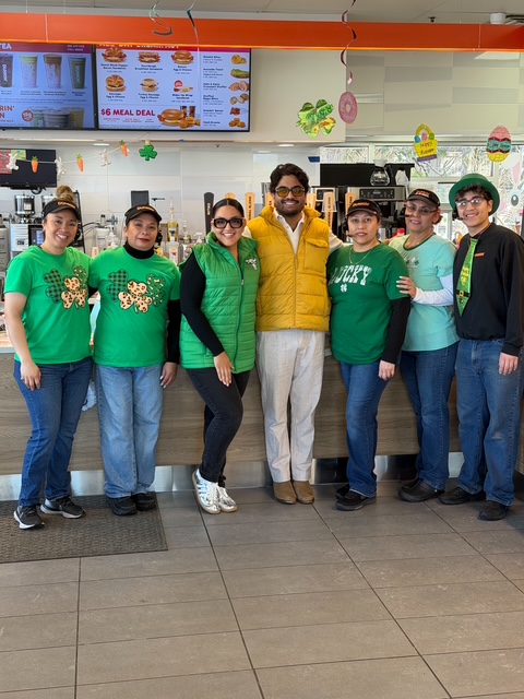 The PPP Donuts Network crew in green, smiling together in front of the counter on St. Patrick's Day